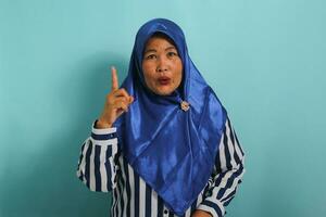 un excité âge moyen asiatique femme, portant une bleu hijab et une rayé chemise, obtient un incroyable idée tandis que permanent contre une bleu Contexte. photo