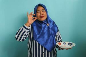 une content âge moyen asiatique femme, portant une bleu hijab et une rayé chemise, est montrant une délicieux geste par embrasser sa les doigts tandis que en portant un vide blanc plaque, permanent contre une bleu Contexte photo