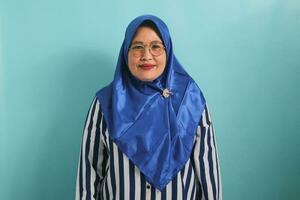 portrait de une âge moyen asiatique femme dans une bleu hijab et rayé chemise, portant lunettes et à la recherche à le caméra. elle est isolé plus de une bleu Contexte photo