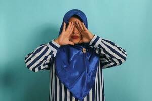 une âge moyen asiatique femme dans une bleu hijab et rayé chemise formes une Triangle avec sa les doigts tandis que permanent contre une bleu Contexte photo