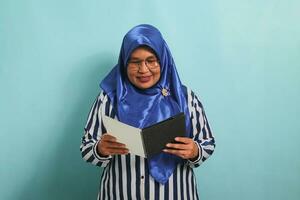 étonné asiatique âge moyen femme, portant une bleu hijab, lunettes, et une rayé chemise, détient un ouvert livre avec une surpris expression tandis que permanent contre une bleu Contexte. photo