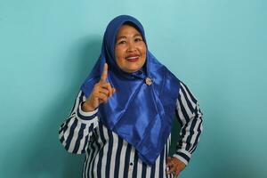 un excité âge moyen asiatique femme, portant une bleu hijab et une rayé chemise, obtient un incroyable idée tandis que permanent contre une bleu Contexte. photo