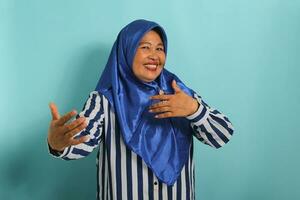 une content âge moyen asiatique femme dans une bleu hijab et rayé chemise étend sa mains vers le caméra, comme si atteindre pour une câlin, isolé contre une bleu Contexte photo