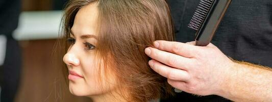 Masculin coiffeur travaux sur le coiffure de le Jeune caucasien brunette femme à une cheveux salon. photo