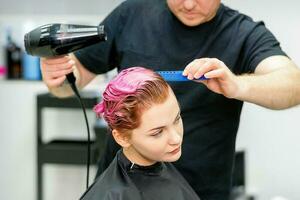une coiffeur est séchage le rose cheveux de le Jeune femme dans une beauté salon. photo