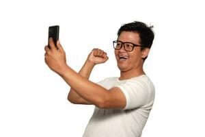 excité asiatique homme avec des lunettes portant blanc T-shirt souriant tandis que en portant le sien téléphone, isolé par blanc Contexte photo