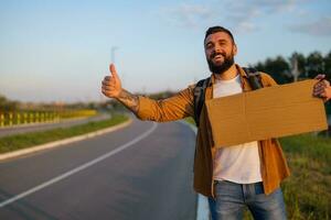 homme est auto-stop sur bord de la route en essayant à Arrêtez auto. il est en portant Vide papier carton pour votre texte. photo