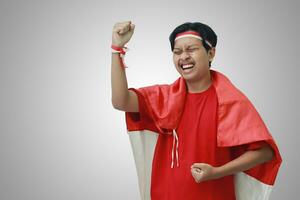 portrait de attrayant asiatique homme dans T-shirt avec rouge blanc ruban sur tête avec drapeau sur le sien épaule comme une manteau, élevage le sien poing, célébrer l'indonésie indépendance journée. isolé image sur gris photo