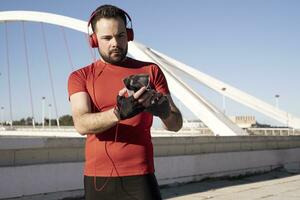 une fermer coup de une Masculin dans rouge écouteurs en utilisant le sien mobile tandis que le jogging dans le rue photo