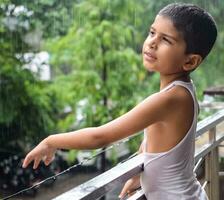 peu enfant en jouant dans été pluie dans maison balcon, Indien intelligent garçon en jouant avec pluie gouttes pendant mousson pluvieux saison, enfant en jouant dans pluie photo