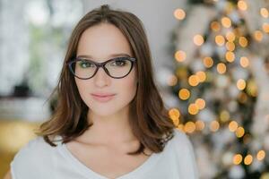 aux cheveux noirs femme dans lunettes, sérieux expression, des stands par Nouveau année arbre à l'intérieur. attirant regarder. photo