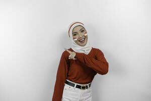 une content asiatique musulman femme portant blanc hijab et rouge haut, et montrer du doigt copie espace sur Haut de son, isolé par blanc Contexte. l'indonésie indépendance journée photo