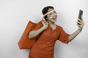 un excité asiatique femme portant rouge kebaya en portant une achats sac et sa téléphone intelligent, studio coup isolé sur blanc Contexte. l'indonésie indépendance journée concept photo