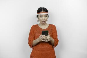 une portrait de une sous le choc asiatique femme portant rouge kebaya et Bandeau, en portant sa téléphone tandis que sa bouche large ouvrir, isolé par blanc Contexte. l'indonésie indépendance journée concept photo