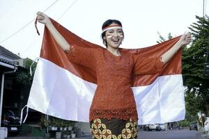content souriant indonésien femme portant rouge kebaya en portant l'indonésie drapeau à célébrer Indonésie indépendance journée. Extérieur séance photo concept