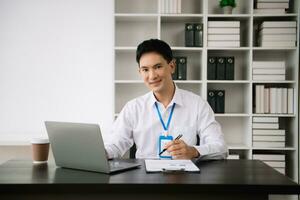 Jeune asiatique affaires homme en utilisant portable et tablette tandis que séance à sa travail lieu. concentré à travail. photo