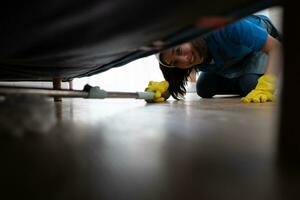 Jeune femme nettoyage le sol avec une balai en dessous de le canapé dans le vivant pièce photo