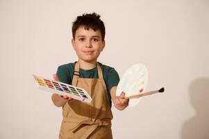 adolescent garçon peintre détient en dehors à caméra une palette avec aquarelles et peindre brosse. Créatif loisir pour enfants. art classe. photo