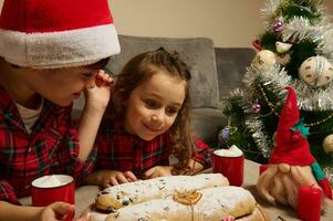 adorable européen enfants, garçon dans Père Noël chapeau et fille, sœur et frère séance à table avec cuit traditionnel allemand volé pain et tasse avec cacao boisson avec guimauves, célébrer Noël photo