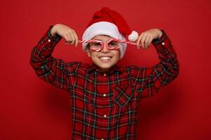 Beau enfant garçon dans Père Noël claus chapeau et plaid chemise sourit à la recherche à caméra par Noël sucré bonbons cannes, en portant leur imitant lunettes, posant sur rouge Contexte avec copie espace pour un d photo