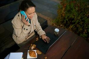 sur de soi aux cheveux noirs femme dans beige tranchée manteau dactylographie sur clavier tandis que parlant sur mobile téléphone. Jeune affaires femme travail sur portable dans Extérieur en bois forêt café après tasse de café photo