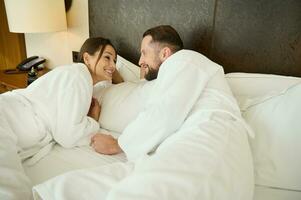 couple dans aimer, jeunes mariés sur leur voyage de noces. magnifique femme portant une blanc éponge peignoir de bain mensonge dans lit avec sa mari et profiter le intimité de le fin de semaine ensemble à maison. famille relation photo