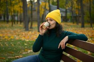 serein Enchanté femme dans chaud de laine Jaune tricoté chapeau séance sur une parc banc et en buvant café de à emporter artisanat papier tasse, repos dans un automnal forêt parc avec érable des arbres sur Contexte photo