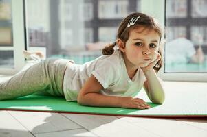 adorable fille relaxant sur un exercice tapis et posant à caméra contre le Contexte de grand les fenêtres à Accueil sur une ensoleillé journée photo