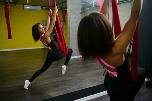 arrière vue à une Jeune svelte corps aptitude femme, africain athlète à la recherche à sa miroir réflexion tandis que pratiquant mouche yoga dans le salle de sport, aérien aéro mouche aptitude. photo