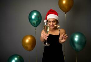 concentrer sur jolie africain femme dans Père Noël chapeau et noir robe célébrer Nouveau année fête avec cierges magiques et Champagne flûte dans mains contre brillant air des ballons sur gris mur Contexte avec copie un d espace photo