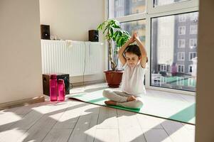 charmant enfant séance sur une aptitude tapis et pratiquant yoga dans lotus position à maison. correct en bonne santé parentalité, méditation photo