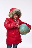 adorable élémentaire vieilli européen garçon dans une chaud couvert de neige rouge vers le bas veste avec une capot, montrer du doigt le sien doigt à le globe dans chercher de une Voyage destination. hiver tourisme concept avec copie un d espace photo