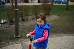 charmant enfant, adorable peu fille équitation pousser scooter dans le ville parc photo