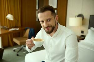 milieu vieilli homme d'affaire, entrepreneur, investissement conseiller dans blanc chemise séance sur lit dans Hôtel chambre pendant affaires voyage, sourit à la recherche à caméra, en portant une tasse avec fraîchement brassée café boisson photo