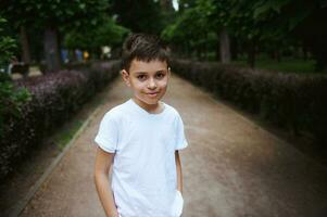 portrait Beau hispanique école âge garçon dans blanc T-shirt, souriant à la recherche à caméra, permanent dans le ruelle de une parc photo