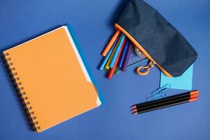 plat allonger avec une cahier, organisateur, coloré des crayons et crayon Cas avec chute en dehors école Provisions , dans bleu et Orange contraste ombres isolé sur bleu Contexte avec copie espace photo