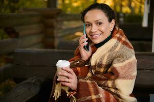 jolie femme regards à caméra parlant sur cellule téléphone, en portant papier carton tasse de chaud boire, en gardant chaud tandis que séance sur en bois banc enveloppé dans une beige confortable à carreaux de laine couverture sur une cool l'automne journée photo