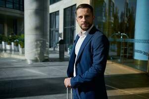 taille longueur portrait de une Beau réussi homme d'affaire développeur entrepreneur dans affaires costume avec une bagage permanent Extérieur sur une entreprise moderne gratte-ciel bâtiment verre entrée Contexte photo