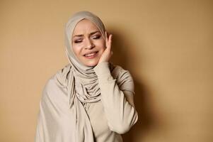 musulman femme dans une strict formel traditionnel robe avec une tête couvert avec une hijab souffre de une mal aux dents. beige Contexte avec copie espace photo