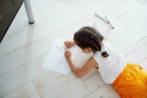 Haut vue de une peu enfant fille mensonge sur le sol à Accueil et dessin image sur une blanc papier feuille à maison. photo