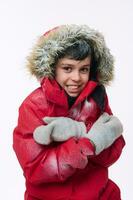 isolé portrait sur blanc Contexte de une Beau européen élémentaire âge garçon portant une brillant rouge veste avec une couvert de neige capuche à la recherche à le caméra, étreindre lui-même, grelottant de le du froid photo