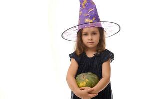 Halloween sorcière concept - à la taille coup de peu caucasien fille habillé dans élégant carnaval tenue et sorcier chapeau, sorcière enfant posant avec citrouille contre une blanc Contexte avec copie espace photo