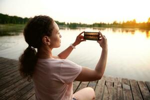 Jeune femme repos sur le jetée et photographier une magnifique le coucher du soleil derrière le Lac sur une été soir photo