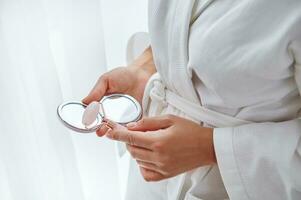 fermer de femelle mains en portant petit cosmétique miroir et jade pierre rouleau masseur. tondu image de une femme dans blanc peignoir de bain avec jade rouleau un une miroir dans sa mains photo