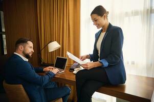 affaires les partenaires travail ensemble dans Hôtel pièce sur affaires voyage. jolie femelle patron avec empiler de papier les documents sourires, en train de lire contact, Beau collègue séance à table et travail sur portable photo