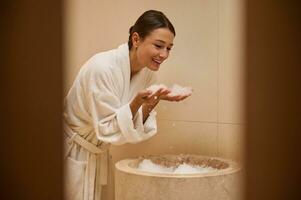 attrayant milieu vieilli aux cheveux noirs caucasien femme habillé dans blanc éponge peignoir de bain repos à bien-être spa, en portant la glace pièces près sa affronter. jeunesse, fraîcheur, corps traitement et des loisirs concept. photo