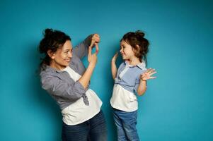 côté vue de une souriant frisé femme à la recherche à sa mignonne bébé fille par sa des doigts, encadrement sa affronter. bleu Contexte avec copie espace photo