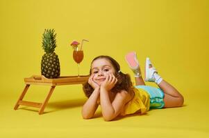 magnifique fille mensonge sur Jaune Contexte près une bambou plateau avec ananas et cocktail dans verre décoré avec parapluie photo