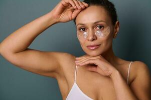 attrayant femme avec anti-fatigue sous les yeux masque à la recherche à caméra posant contre gris Contexte. peau se soucier, hydrogel collagène patchs, en tissu masque en dessous de yeux à réduire œil Sacs et gonflement photo