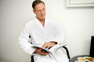 Beau européen milieu vieilli affaires homme portant blanc éponge peignoir de bain, séance sur un fauteuil et en train de lire magazine tandis que repos dans Hôtel pièce de bien-être spa centre photo